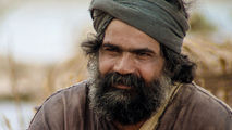 Man with a beard and headscarf looks contemplative outdoors. He wears a brown robe, set against a blurred natural background.