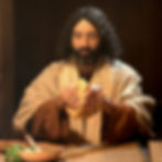 Man with long hair breaking bread at a table. Warm lighting, earthy tones, and a rustic setting. Calm and focused expression.