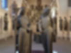 Two large, weathered statues in a museum, one holding a cross and chalice. Other smaller sculptures and archways in the background. Warm lighting.