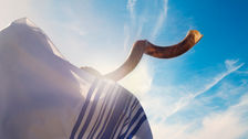 A shofar held up against a bright blue sky, with a striped white fabric partially visible, conveying a serene and spiritual mood.
