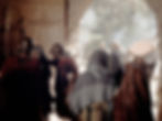 People in historical attire congregate near an arched doorway. Roman soldiers with helmets stand by, while others in hooded robes exit into sunlight.