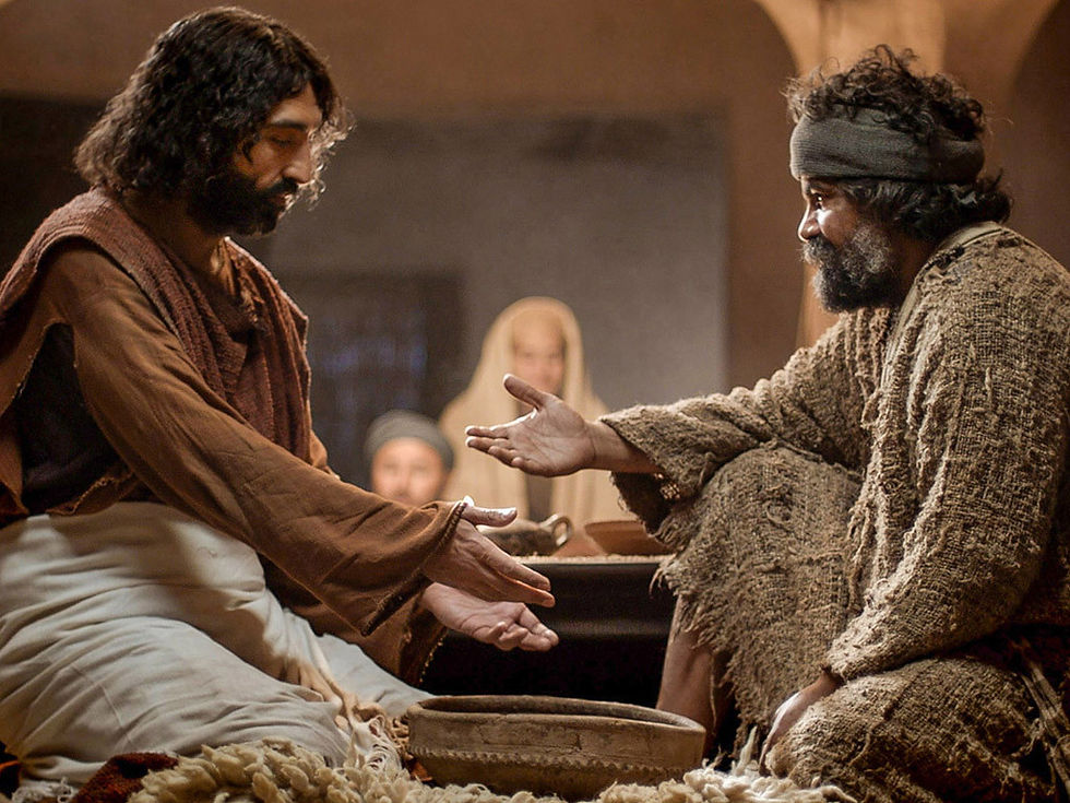 Two men in robes sit indoors, gesturing toward each other with open hands. A bowl is between them. Soft lighting creates a peaceful mood.