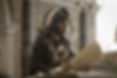 Man in ornate medieval robe and hat reads a large open book in a stone-walled room. The mood is serious and focused.