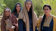 Four women in traditional clothing stand outdoors, looking intently. One holds a clay jug. Trees and mountains in the background.