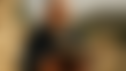 Bald man with glasses reads a book outdoors in a sunny, ancient ruin setting, with stone walls and hills in the background, focused mood.