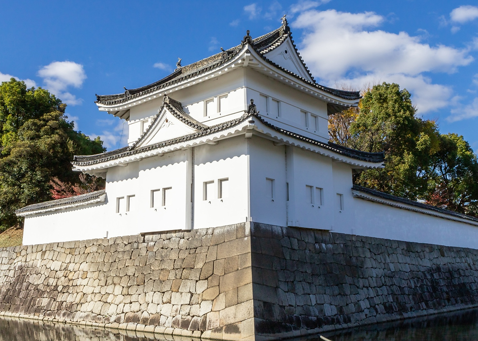 White Japanese castle turret against a blue sky, surrounded by green trees. Stone wall and water in the foreground. Peaceful setting.