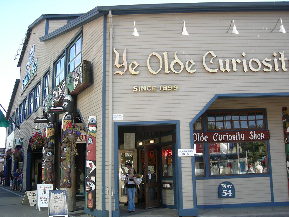 Pier 54 Olde Curiosity Shop storefront with totem poles, a woman exiting, and an OPEN neon sign on a sunny pier.