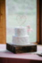 Two-tier white cake with textured icing on a wooden stand. A wire heart topper adorns the cake. Background shows blurred greenery inside a wedding venue near Morehead City NC. Eastern NC wedding venue. Affordable wedding venue.