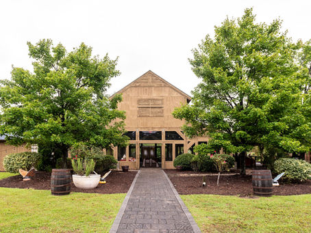 Butterfly Kisses Pavilion- Wooden barn-style building framed by leafy trees and shrubs, with a stone path leading to the entrance on a bright overcast day.