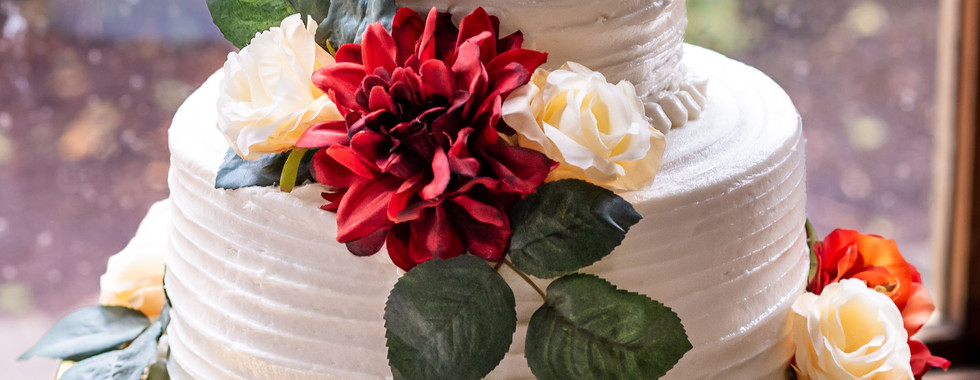 a two tiered white wedding cake with flowers and a Mr. and Mrs. cake topper. The cake sits on a gold cake stand. The background is a window that looks into a landscaped wedding venue in Newport, NC.