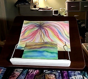 Portable art workspace set up beside a bed with colorful watercolor paintings and painting supplies within easy reach.