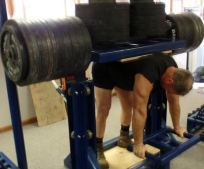A figure bent over in a gym machine, with a loaded platform/ bar on their back. There are dozens of weight plates, weighing hundreds of kilos on the bars.