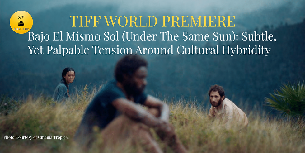 Three people sit in a grassy field on a mountainside, with misty blue mountains in the background. One person in the foreground is out of focus, while a woman on the left and a bearded man on the right are in focus, both looking toward the camera with solemn expressions.