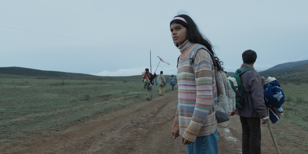 A girl in a striped sweater stands on a dirt road with a backpack. People walk in the distance on a cloudy day, evoking a somber mood.
