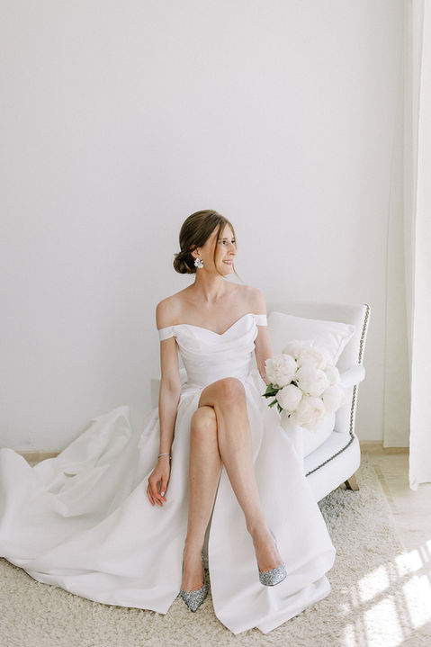 Portrait éditorial de la mariée assise dans sa robe de mariage, chaussures et bouquet de fleurs.