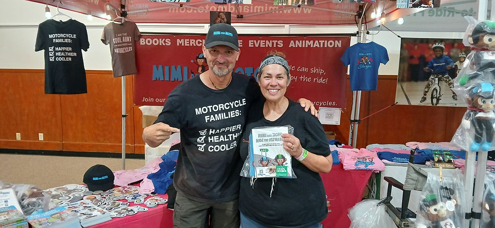 Mark Augustyn pointing to his Motorcycle Families shirt beside a rider holding Mimi and Moto Ride the Alphabet at an indoor event booth.