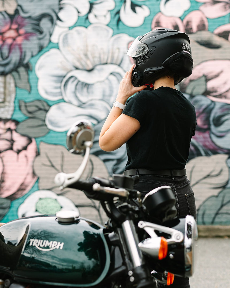 Jen putting on her Shoei helmet with a Hightail hair protector beside her Triumph Thruxton in front of a floral mural.