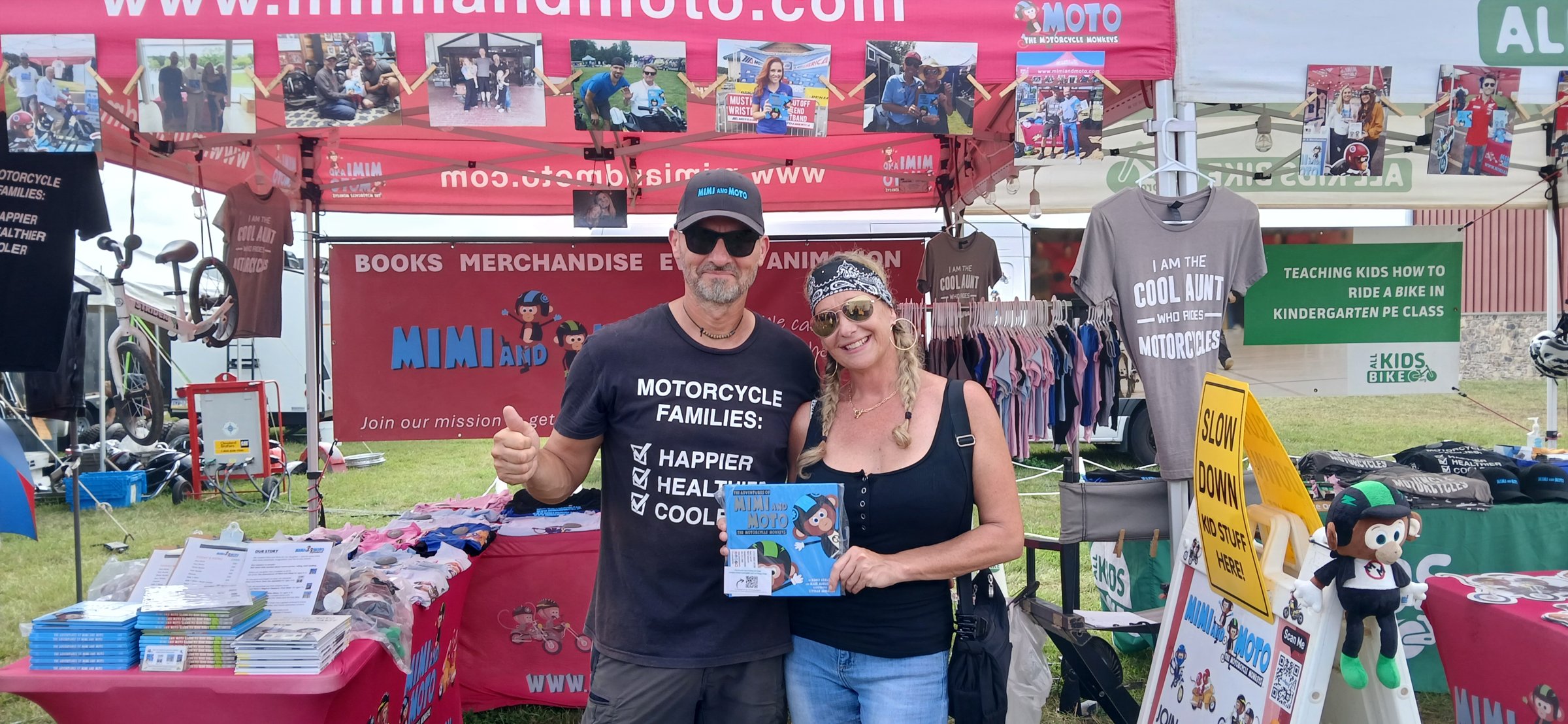 Mark Augustyn of Mimi and Moto giving a thumbs up beside a rider holding a Mimi and Moto book at their booth at AMA Vintage Motorcycle Days.