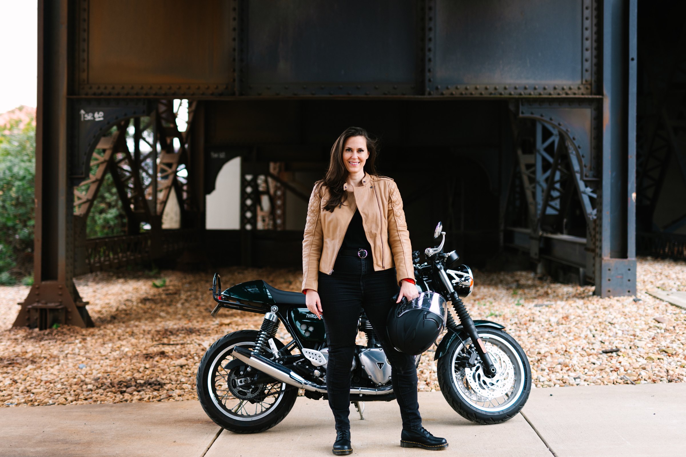 Jen, founder of Hightail Hair, standing in front of her green Triumph Thruxton under a riveted steel rail bridge, holding a Shoei helmet.
