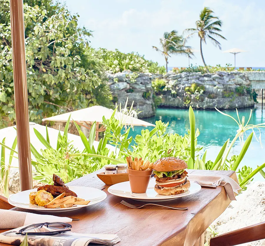 Beachfront dining table with gourmet burger, fries, and oceanfront lagoon views at Hotel Xcaret Mexico, highlighting scenic outdoor dining, tropical surroundings, and all-inclusive resort experience in Riviera Maya.
