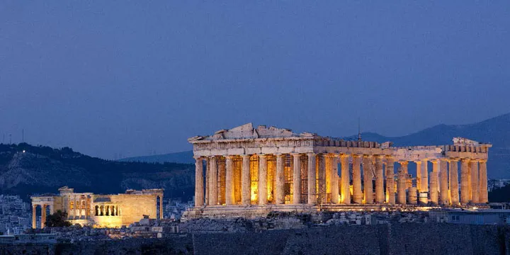 The illuminated Parthenon atop the Acropolis in Athens, Greece, glows against a deep blue evening sky with city lights twinkling below. The ancient temple’s golden columns highlight the timeless beauty and history featured on many Viking Ocean Cruise itineraries.
