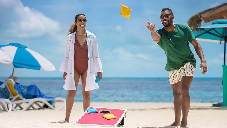 A couple playing cornhole on a sunny beach, with a man tossing a yellow beanbag toward a pink game board while a woman watches and smiles. Beach loungers and umbrellas sit behind them, and the calm blue ocean and clear sky create a relaxed, tropical atmosphere.