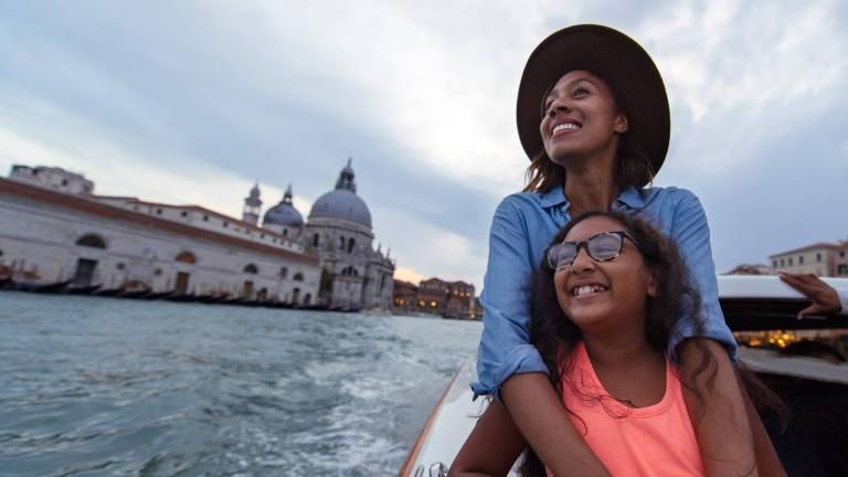 Mother and daughter enjoying a private boat ride along the canals of Venice, Italy with iconic basilica in the background, experiencing an Adventures by Disney European family vacation featured in March 2026 promotions planned by Disney travel expert Brandi Parker.