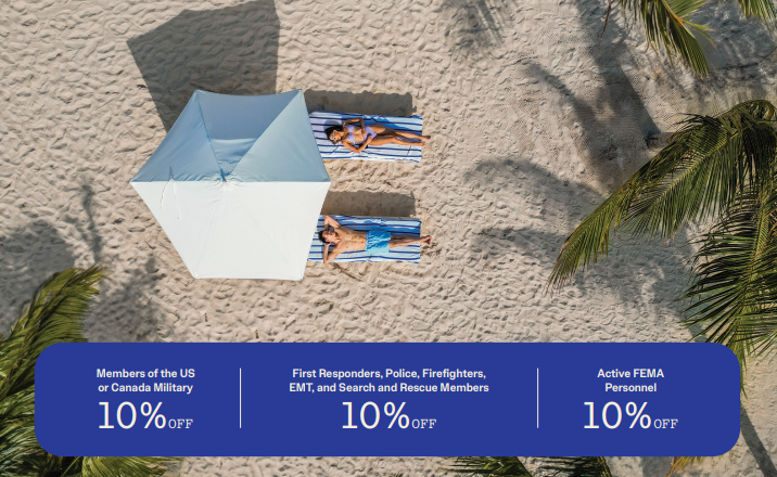 “Overhead view of a couple relaxing on lounge chairs beneath a white beach umbrella on a sandy shoreline surrounded by palm tree shadows at a Sandals Resort. Promotional banner along the bottom highlights 10% off for U.S. or Canada military members, first responders, police, firefighters, EMTs, search and rescue members, and active FEMA personnel. © Sandals Resorts.”