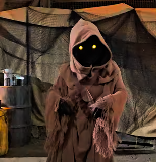 A small robed Jawa character from Star Wars stands with glowing yellow eyes visible beneath a brown hood, holding its hands together in a desert-themed setting with crates and fabric tents in the background.