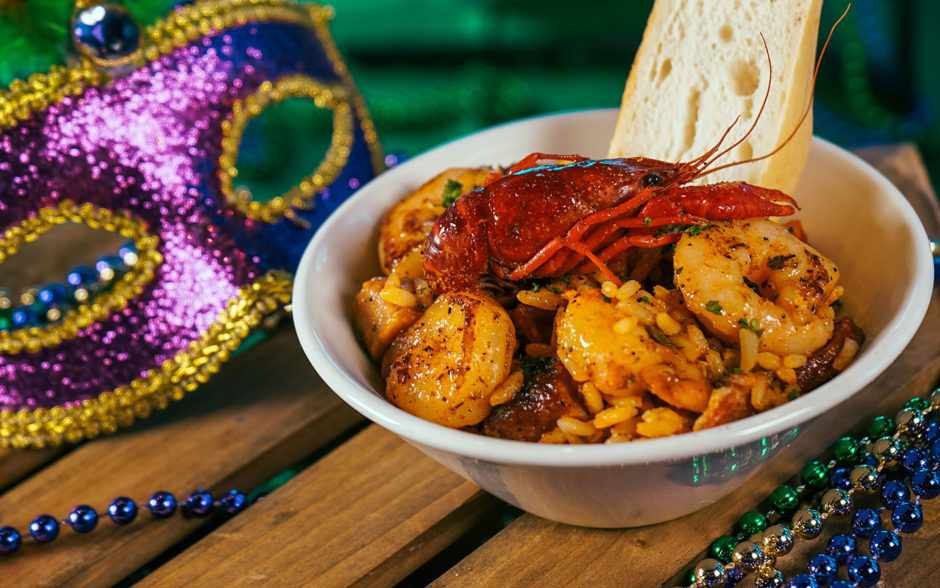 Mardi Gras–inspired Cajun seafood dish featuring shrimp, crawfish, and seasoned rice served in a bowl with toasted bread, surrounded by colorful beads and a decorative mask at Universal Orlando’s annual Mardi Gras celebration.