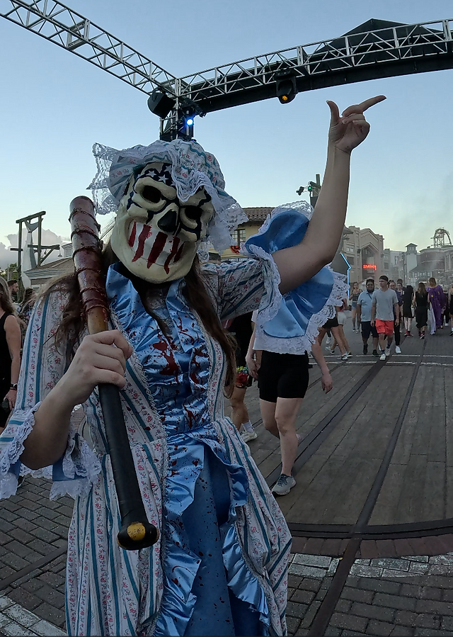 Halloween Horror Nights scare actor in grotesque mask interacting with guests in an outdoor scare zone along a themed boardwalk setting.