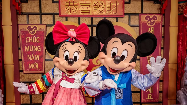 Mickey Mouse and Minnie Mouse posing together in colorful traditional Lunar New Year outfits, standing in front of festive red and gold décor with cultural banners and symbols, celebrating Lunar New Year at Disneyland Resort.