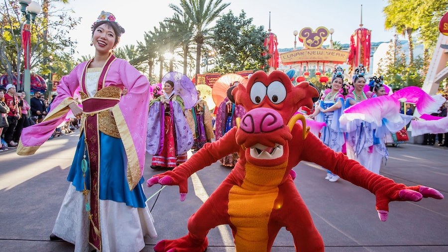 Disneyland Lunar New Year parade scene featuring Mushu dancing in the foreground alongside performers in colorful traditional attire, with festive banners, lanterns, and cultural costumes celebrating Lunar New Year at Disneyland Resort.
