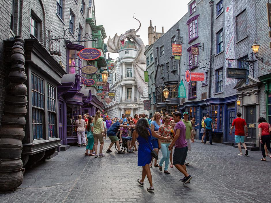Guests stroll through Diagon Alley at Universal Studios Florida, with whimsical storefronts, hanging shop signs, and the towering dragon atop Gringotts Bank in the distance, highlighting immersive Wizarding World theming and a must-see Universal Orlando park experience.