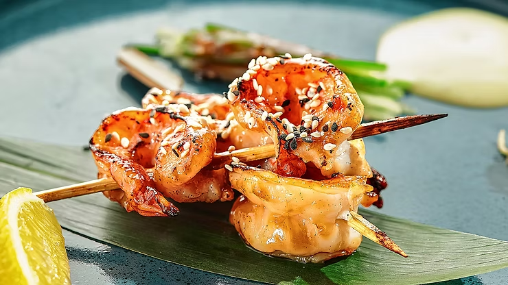 Grilled shrimp skewers glazed in a glossy sauce and topped with sesame seeds, served on a dark plate with a green leaf underneath. A lemon wedge and fresh green garnish sit nearby, highlighting a plated, restaurant-style presentation with warm lighting and crisp detail.