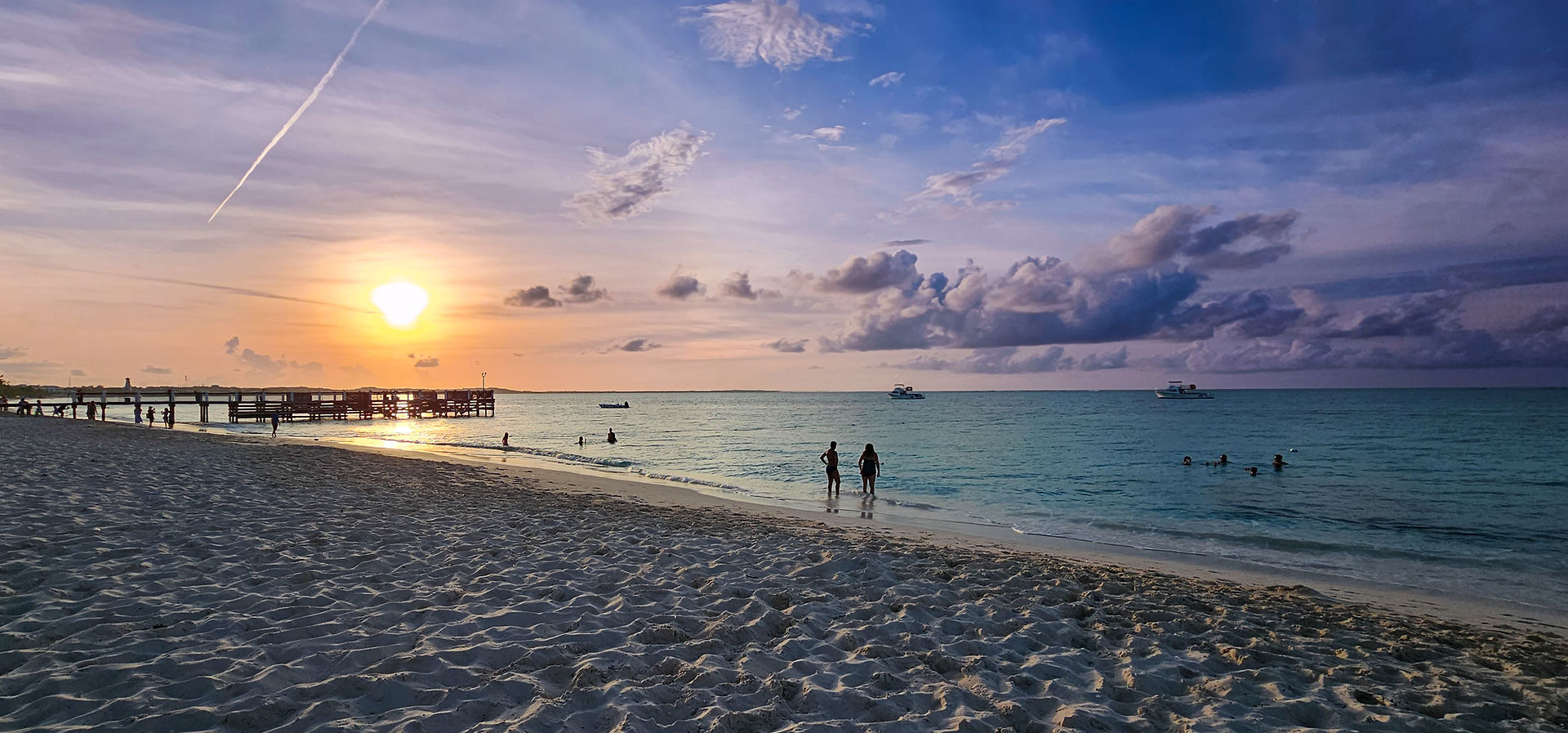 Beaches Turks & Caicos beach at sunset with two people walking along the shoreline, calm turquoise water, and pastel sky, showcasing romantic Caribbean scenery and family-friendly all-inclusive vacation experience.