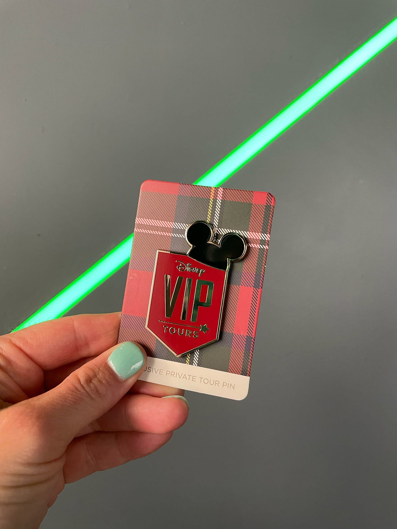 Hand holding a Disney VIP Tour credential badge with Mickey ears design and a green lightsaber in the background, symbolizing exclusive access and the personalized magic of a Walt Disney World VIP experience.