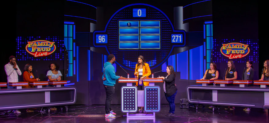 Carnival Cruise Line Family Feud Live game show experience onboard a cruise ship, featuring teams competing on a brightly lit stage with scoreboards, podiums, and live interactive entertainment for families and groups at sea.