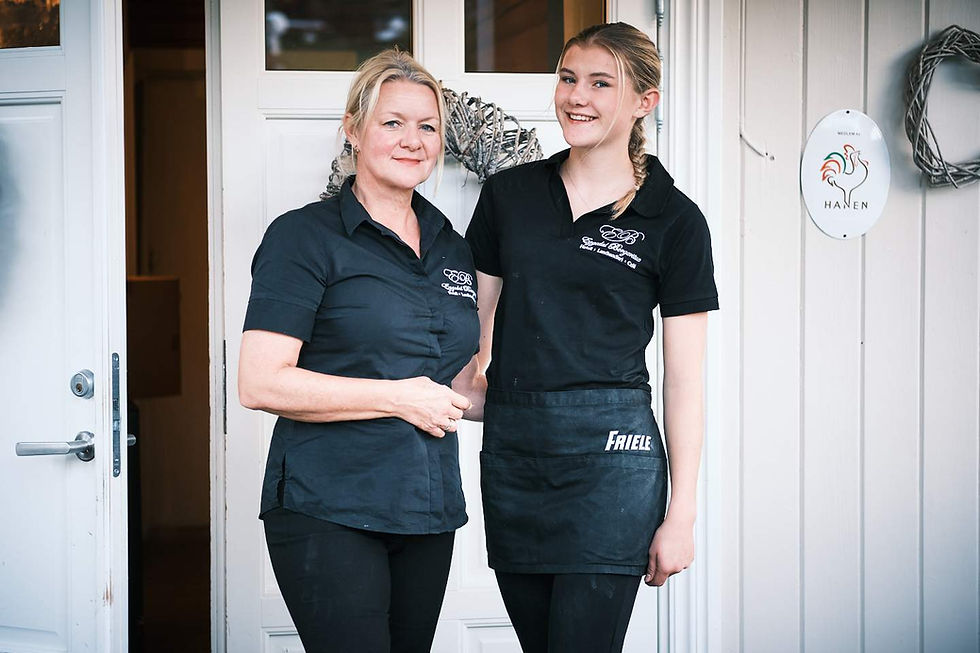 Two women in black uniforms stand smiling at a doorway. Light wood siding and a "Hanen" sign are visible in the background.