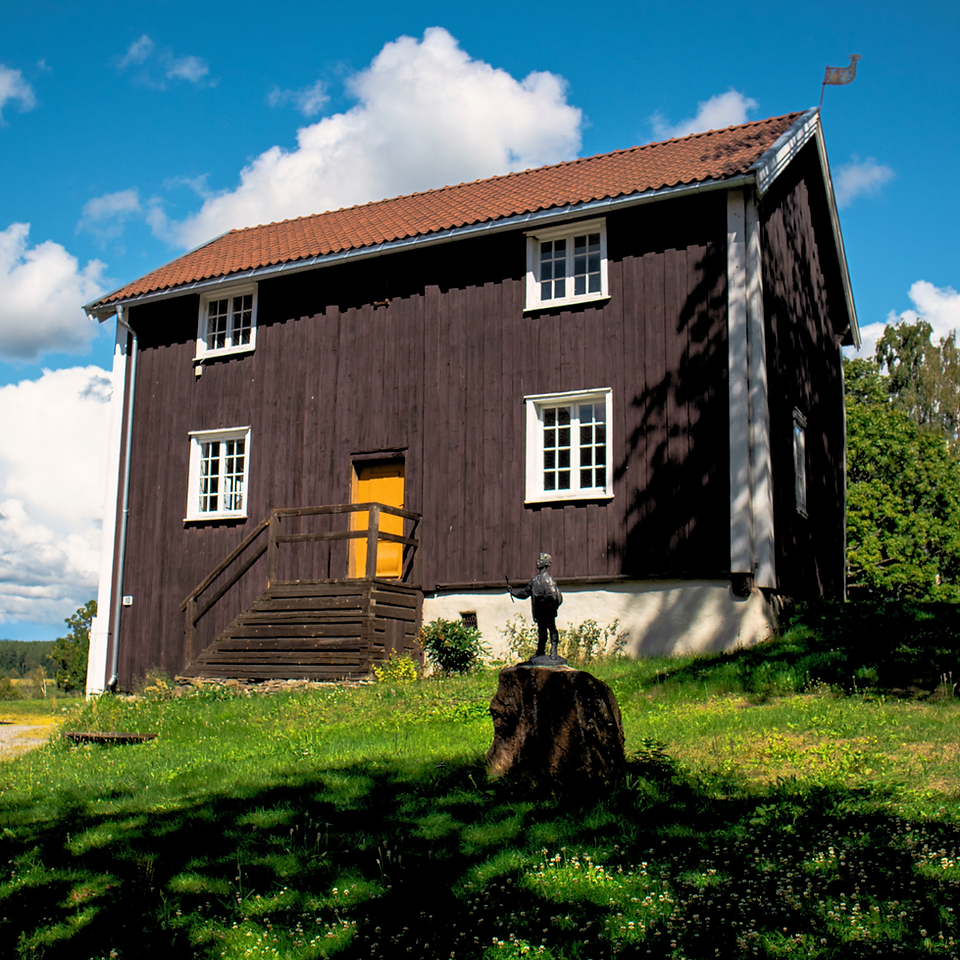 The Old Monk House at Ringerikes Museum standing tall with a yellow door. The building is from the early 18th century and is painted brown with white windows. The sky is blue and the building is surrounded by green trees and grass. 