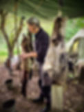 Woman working with animal hides, outdoor camp setting, tree branches, tent backdrop.