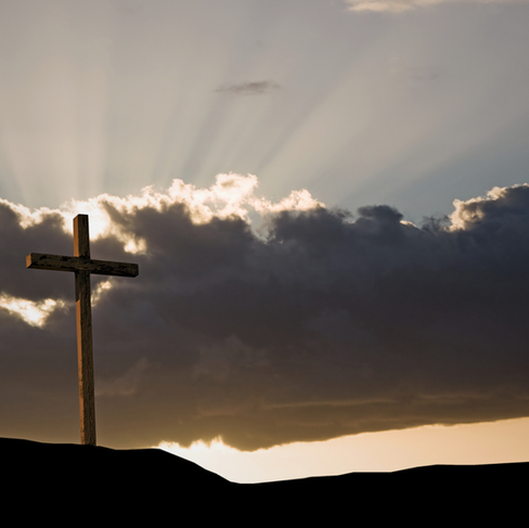 A tall wooden cross stands on a hill underneath puffy clouds with sunrays peering out.