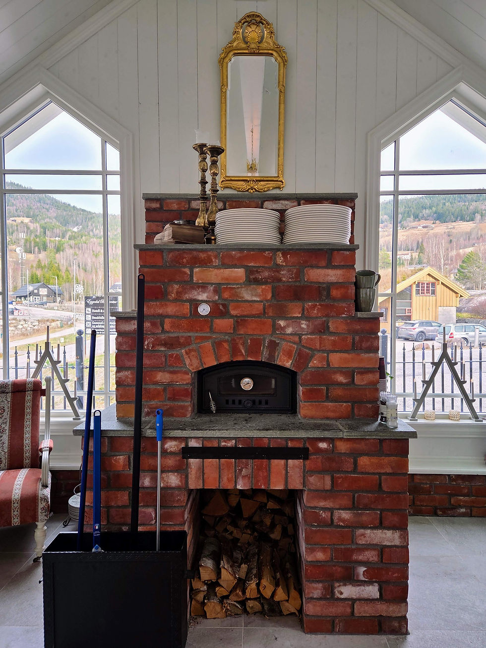 Brick oven with stack of plates, candles, and mirror above. Firewood stored below. Large windows show a scenic mountain view. Cozy setting.