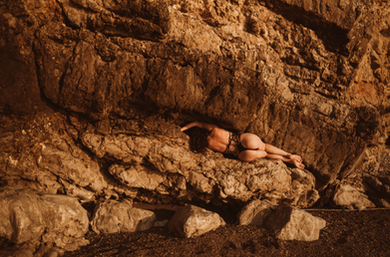 A woman in black couture lingerie layed on a rocky ledge blending with the warm rock wall in soft sunlight. 
