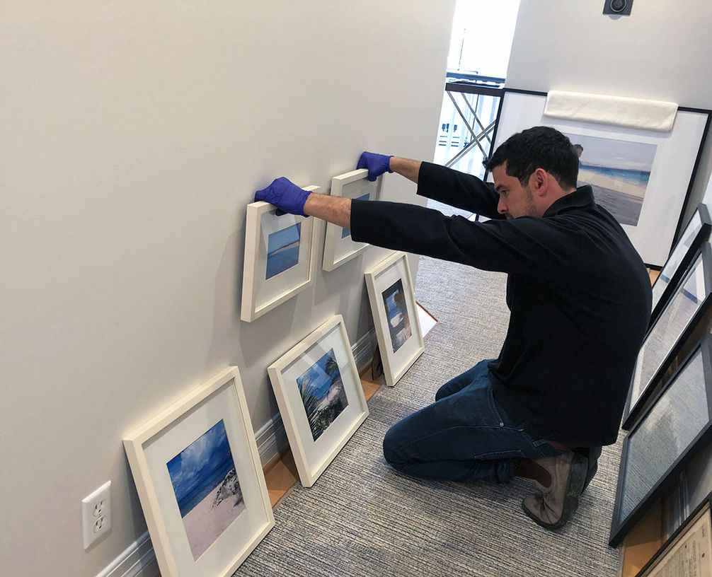 Benjamin Milch - Secure Installations LLC art installer - holds two small framed artworks against the wall to assess their placement.  Three more similar artworks lay against the wall on the floor.  