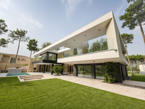 Modern house, white plaster, grey concrete, dark-brown wood, glass windows and doors, glass railings, swimming pool, olive tree, lawn, pine trees, landscaping rocks, beige paving stones, spring, daytime sun.