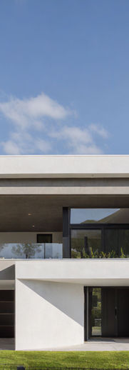 Modern house, white stucco, gray concrete, dark brown composite panels, glass railings, large windows, recessed lighting, flat roof, pine trees, manicured lawn, security fence, paved driveway,  clear sky, daytime sunlight.
