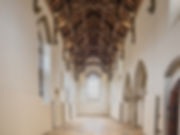 Inside a medieval chapel which is long and narrow. The walls are smooth and painted off-white, there are windows on the sides and a large leaded window at the far end, in the centre of the image. There is a timber roof and a tiled floor.a 