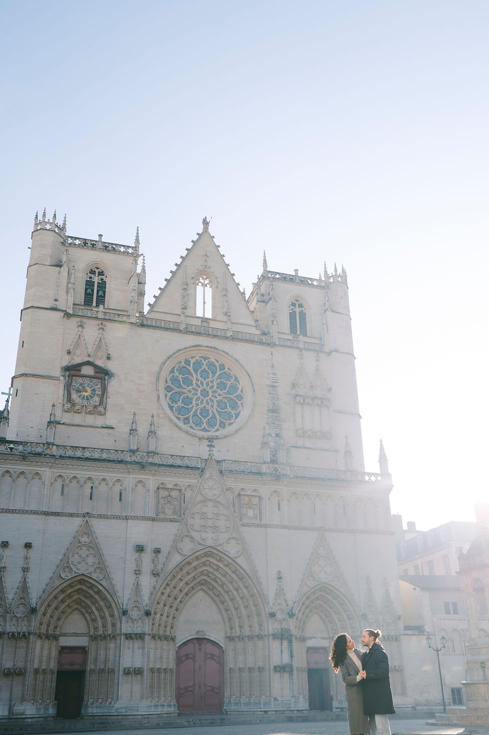Un couple se tient devant une cathédrale gothique Saint-Jean à Vieux-Lyon avec une grande rosace, baigné par la lumière du matin, créant une ambiance romantique.
