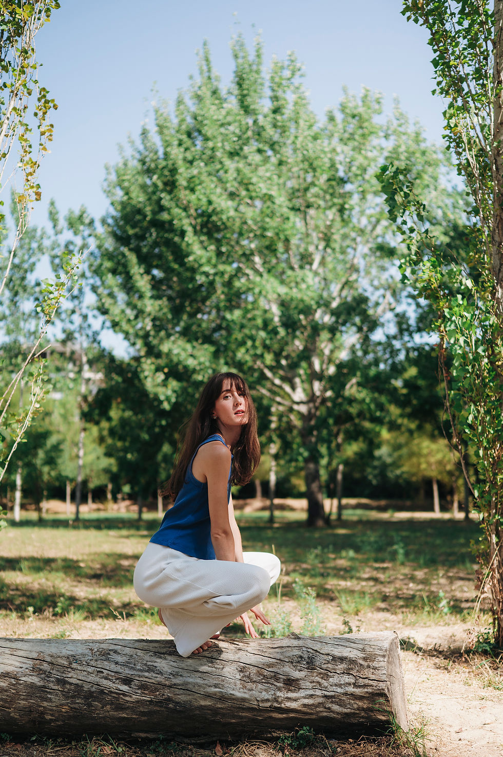 Jeune femme en haut bleu et pantalon blanc, accroupie sur un tronc dans un parc ensoleillé, entourée d’arbres verts.